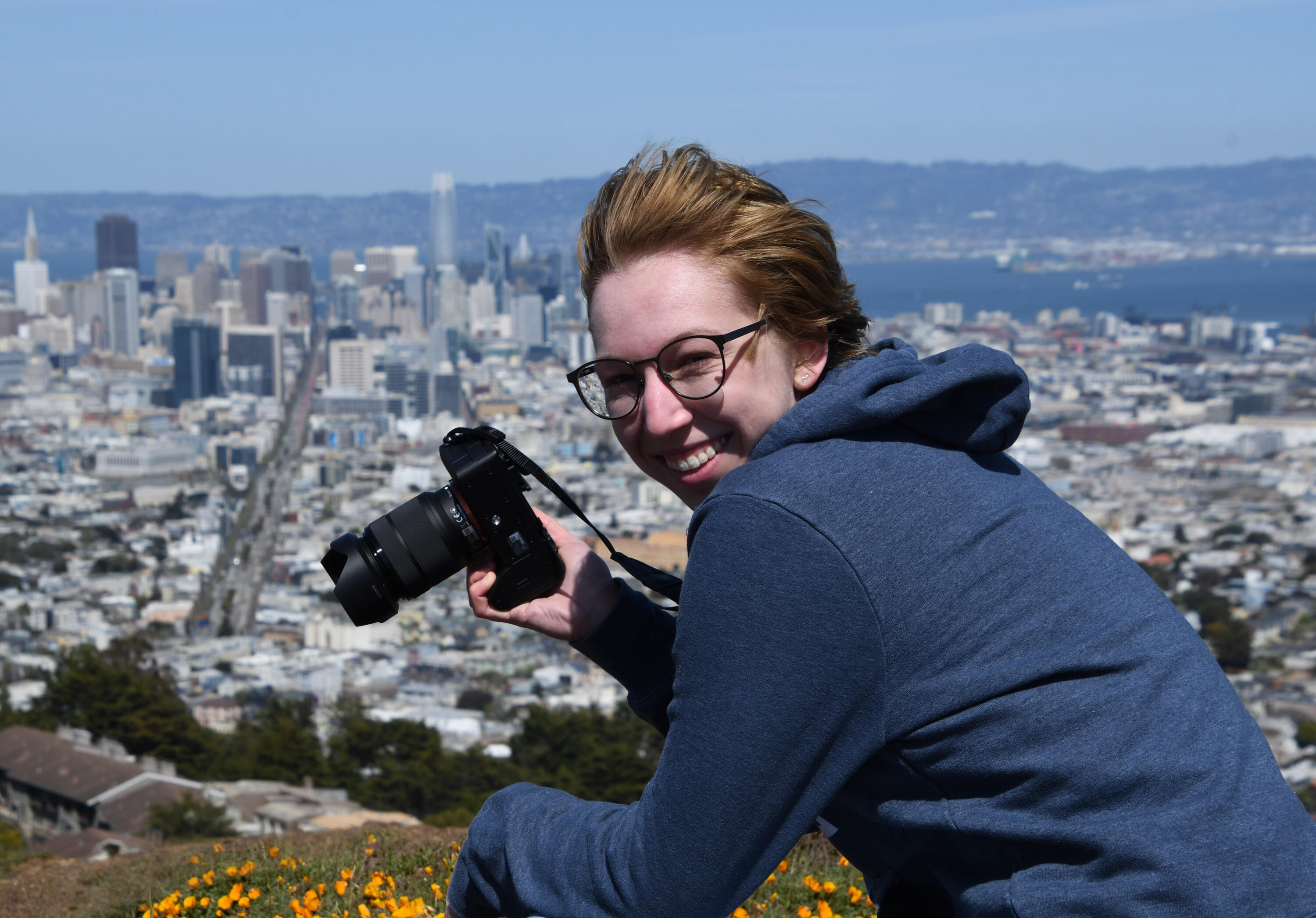 Natalie Gospe in their Element A photo shows a smiling Natalie Gospe as the hold a camera to take photos over the San Francisco skyline. There are California Poppies in bloom below them.